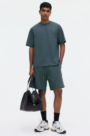 Man wearing dark green t-shirt and shorts holding a black bag against white background
