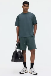 Man wearing dark green t-shirt and shorts holding a black bag against white background