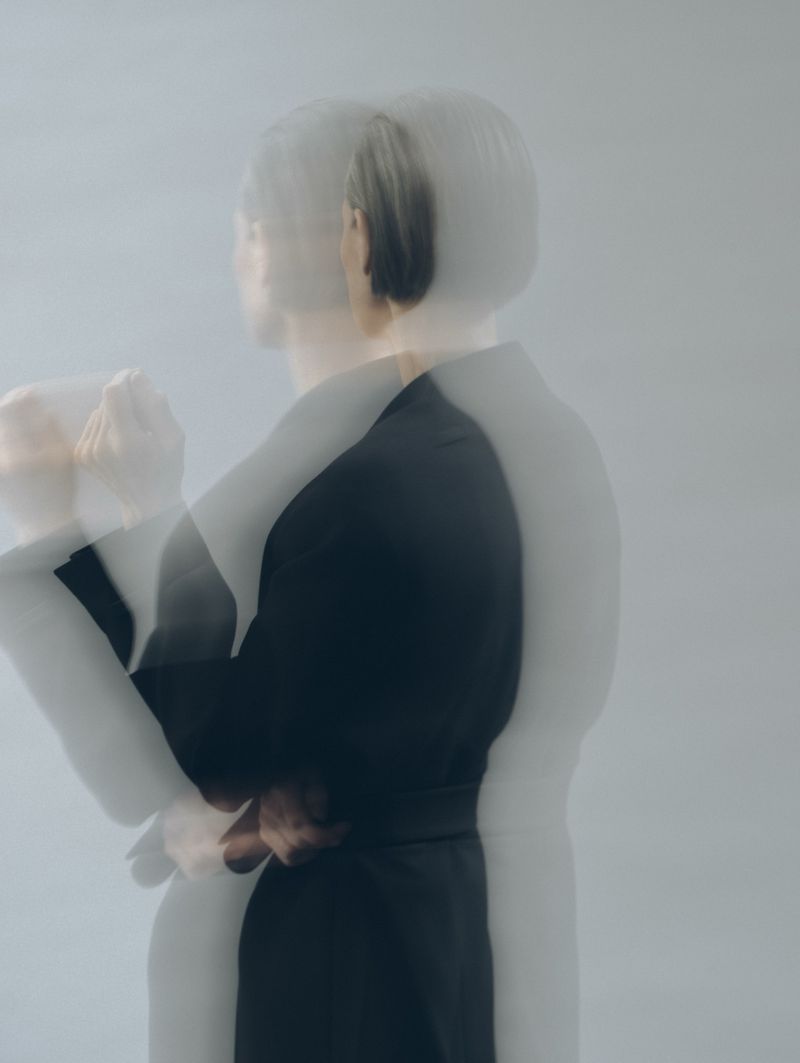 Abstract studio portrait of a woman with multiple exposures in a black top against a muted gray background, creating an ethereal, blurred effect.