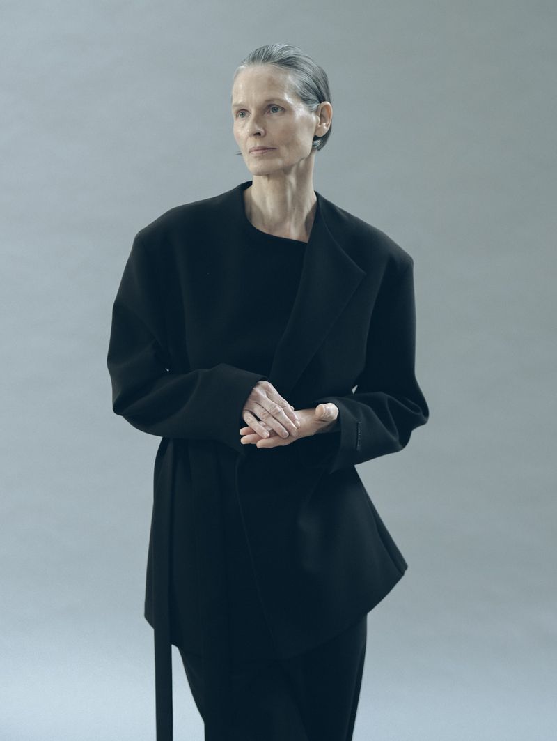 Elegant mature woman in a black belted jacket and skirt poses in a minimalist studio setting, creating a sophisticated and timeless fashion image.