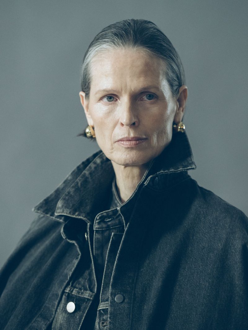 Portrait of an older woman with short grey hair wearing denim jacket and gold earrings against a solid grey backdrop in studio light.