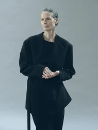 Model wearing a black tailored jacket with clasped hands against a neutral grey studio backdrop.