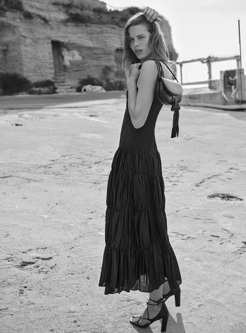 Monochrome full shot of an adult woman posing outdoors wearing a long dress, sandals, and a shoulder bag against a stone cliff backdrop.