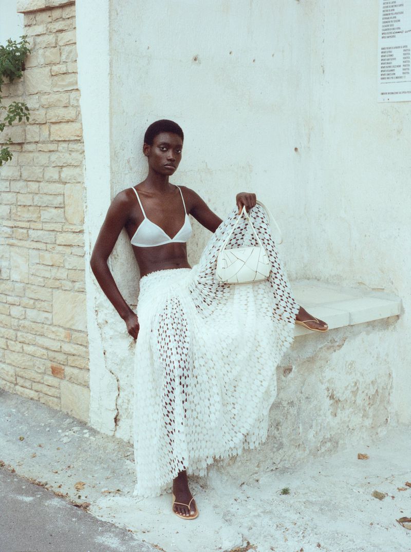 An adult female model with a bra top and a skirt is sitting and posing in an outdoor fashion shot near a wall, holding a handbag.