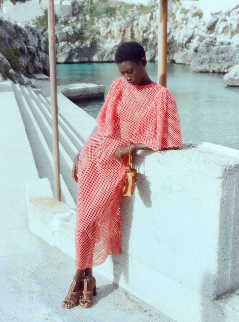 An African American woman in a peach-colored dress sits on a concrete wall with a purse; a clear blue ocean is visible in the background.
