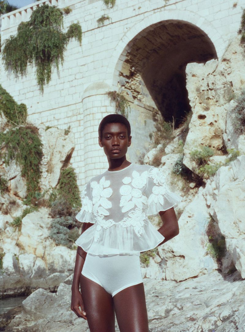 Elegant woman in floral top and briefs poses against a stone bridge and rocky coastline. Fashion, beauty, and coastal vibes.