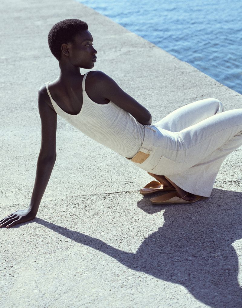 An elegant African-American woman poses gracefully on a concrete platform by the ocean in cream-colored pants and top, wearing flat shoes.