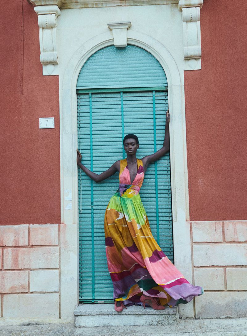 A tall, slim woman poses in a colorful dress against a textured red building and teal shuttered window, creating a vivid fashion portrait.