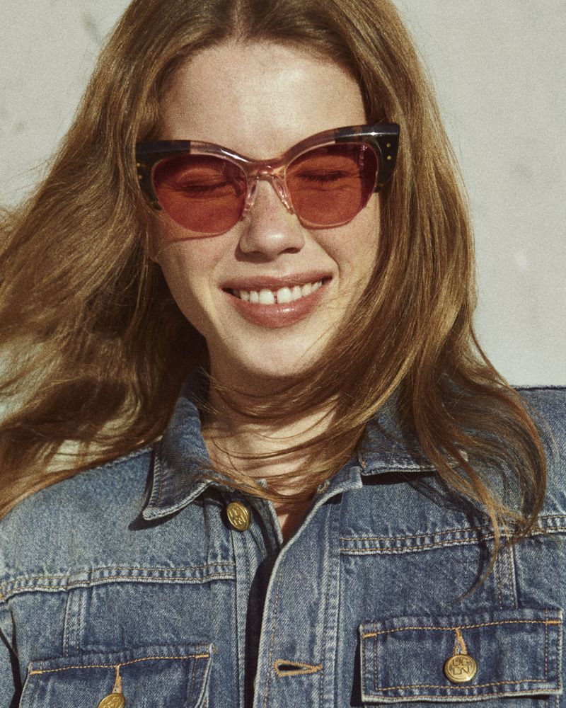 A smiling woman with red sunglasses and a denim jacket gazes directly at the camera in a well-lit, close-up portrait.