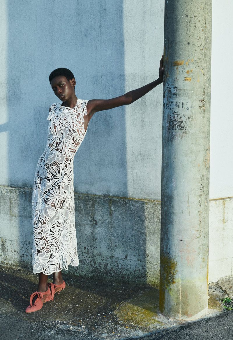 Elegant black woman in a white lace dress poses outdoors, leaning against a weathered pillar in soft, natural light.
