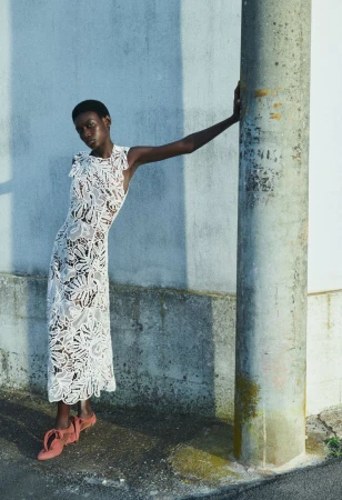 Elegant black woman in a white lace dress poses outdoors, leaning against a weathered pillar in soft, natural light.
