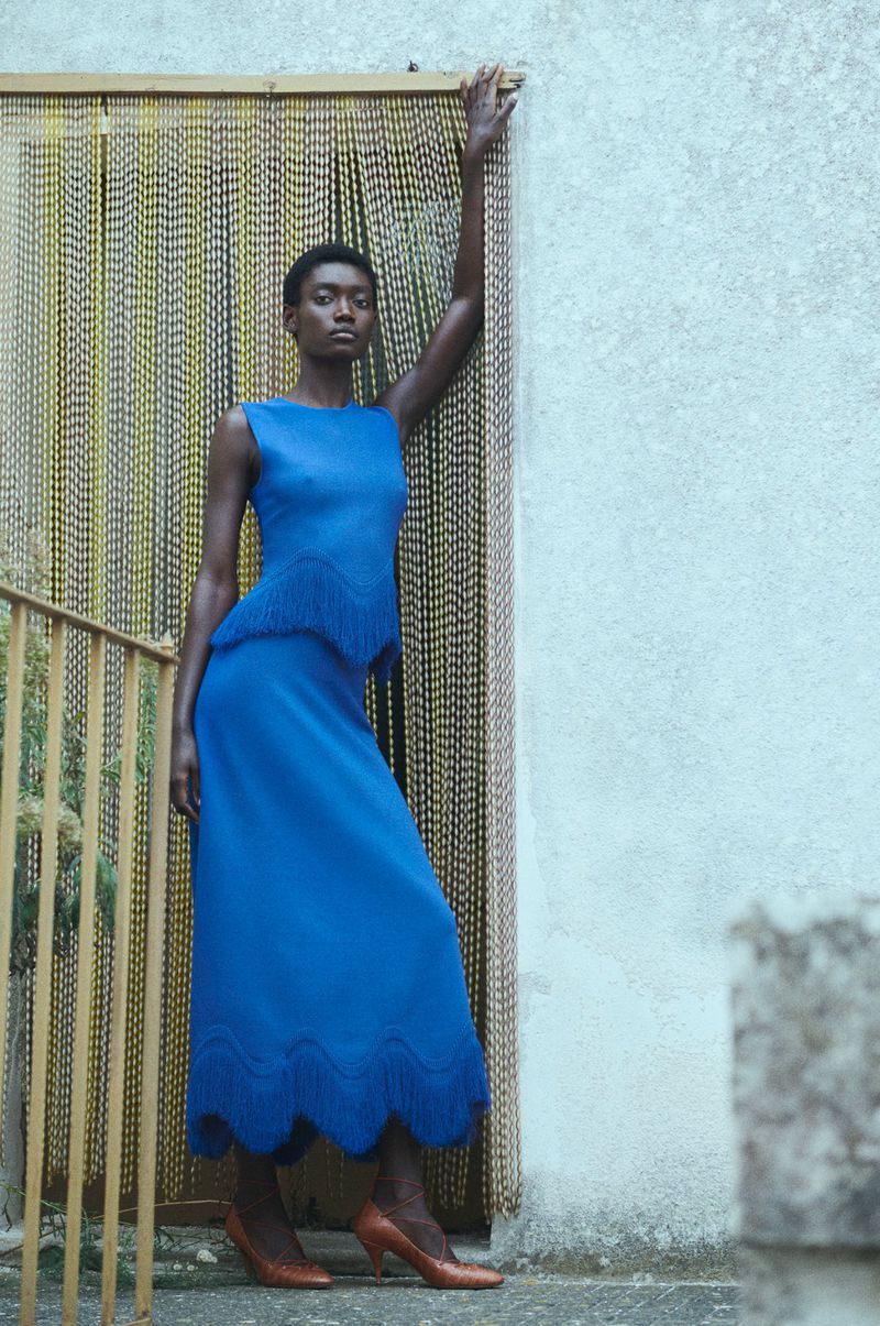 An African woman poses outdoors wearing a blue top and skirt, leaning against a wall near a beaded curtain and stairs, fashion shot.