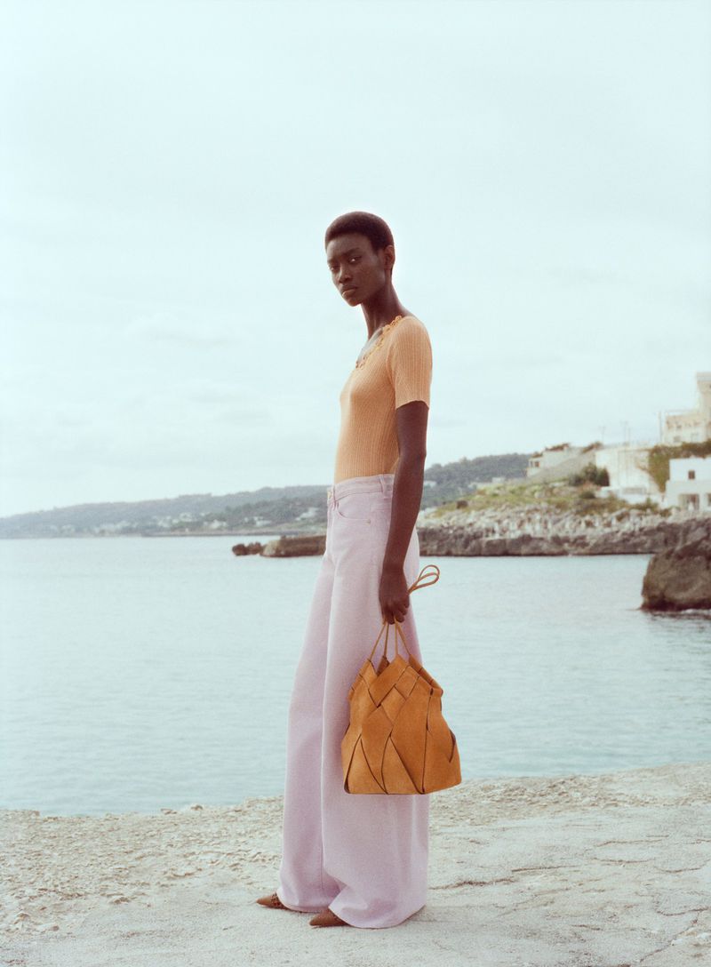 An adult woman stands on a stone surface holding a purse with a blue ocean and cloudy sky in the background, wearing pink pants.