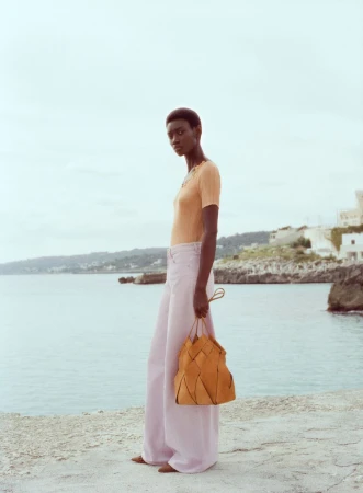 An adult woman stands on a stone surface holding a purse with a blue ocean and cloudy sky in the background, wearing pink pants.