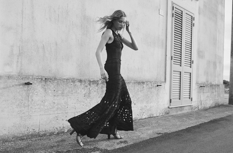 A lone woman in a black dress walks along a grey pavement beside a white wall with a closed shuttered door in the background.