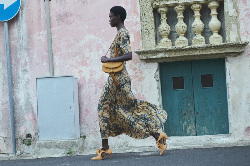 Fashion model in patterned dress and high heels, walking outdoors with a crossbody bag. Pink building and teal door in background.