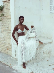 Model in a white crochet top and skirt holding a white handbag against a textured stone wall