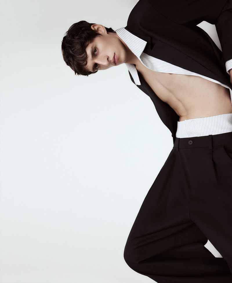 Fashionable young man poses in unbuttoned black suit jacket and wide-legged trousers against a plain white studio backdrop.