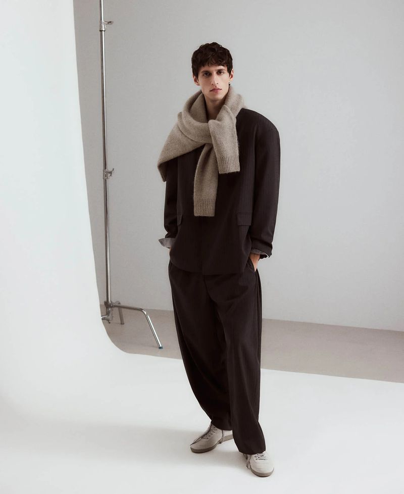 Adult male stands in photo studio wearing oversized black shirt and pants, grey scarf, and light grey sneakers against white background.