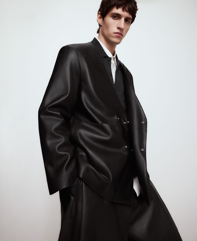 Man poses in a black leather blazer and pants against a white backdrop, showcasing a modern, stylish, and edgy fashion statement.