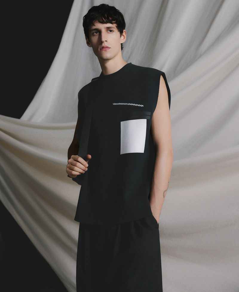 Male model poses in black sleeveless shirt and long skirt against draped white fabric backdrop in studio. Fashion editorial style portrait.