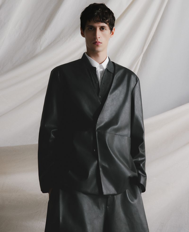 Young man in a black leather-like blazer and pants posing in a studio setting with a neutral-toned backdrop, fashion-forward, cool expression.