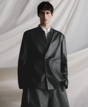 Young man in a black leather-like blazer and pants posing in a studio setting with a neutral-toned backdrop, fashion-forward, cool expression.
