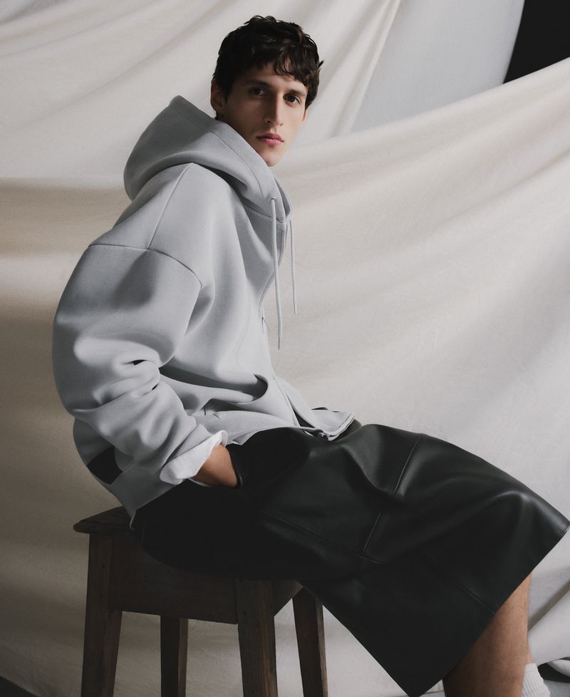 Young man in a light grey zip-up hoodie and black leather shorts sitting on a wooden stool against a draped fabric background.