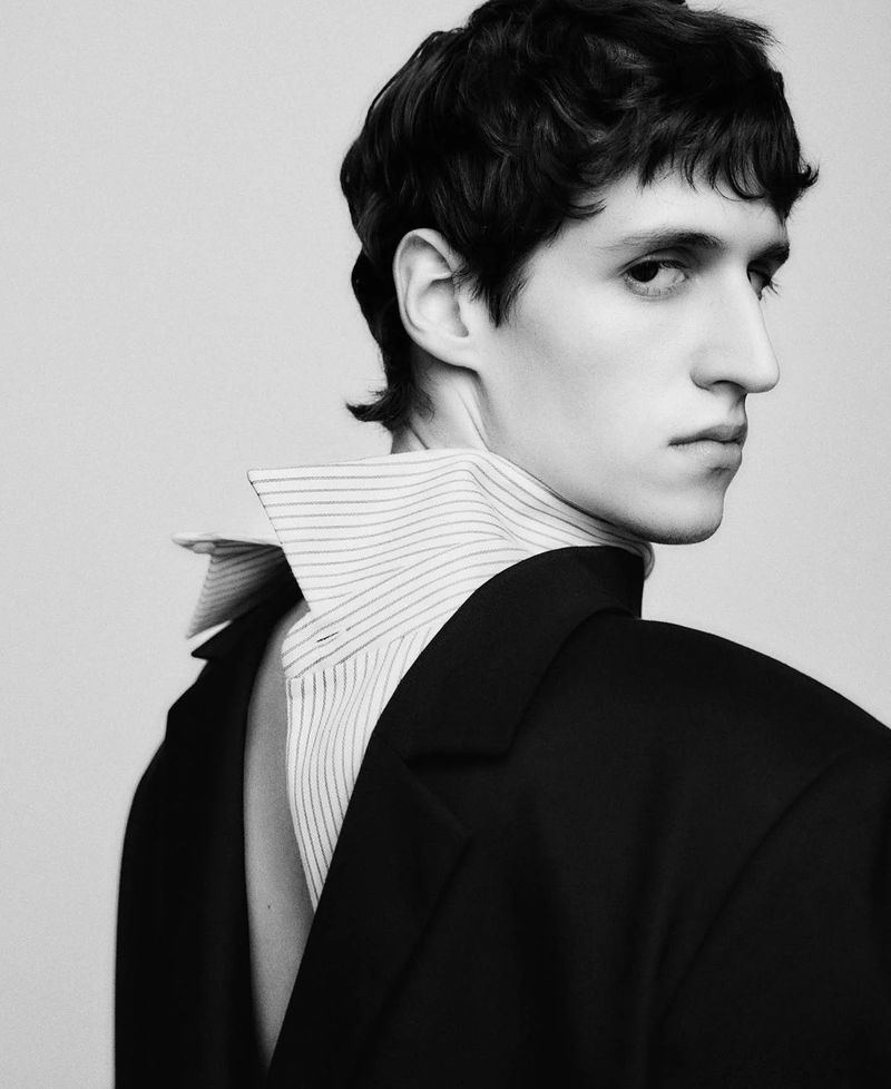 Monochrome portrait of a male model wearing a black jacket backward with a striped collared shirt against a light grey background.