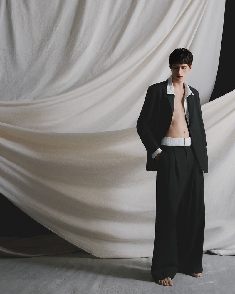A shirtless man wearing a black suit jacket and wide-legged pants poses in front of a draped white fabric backdrop in a fashion editorial.