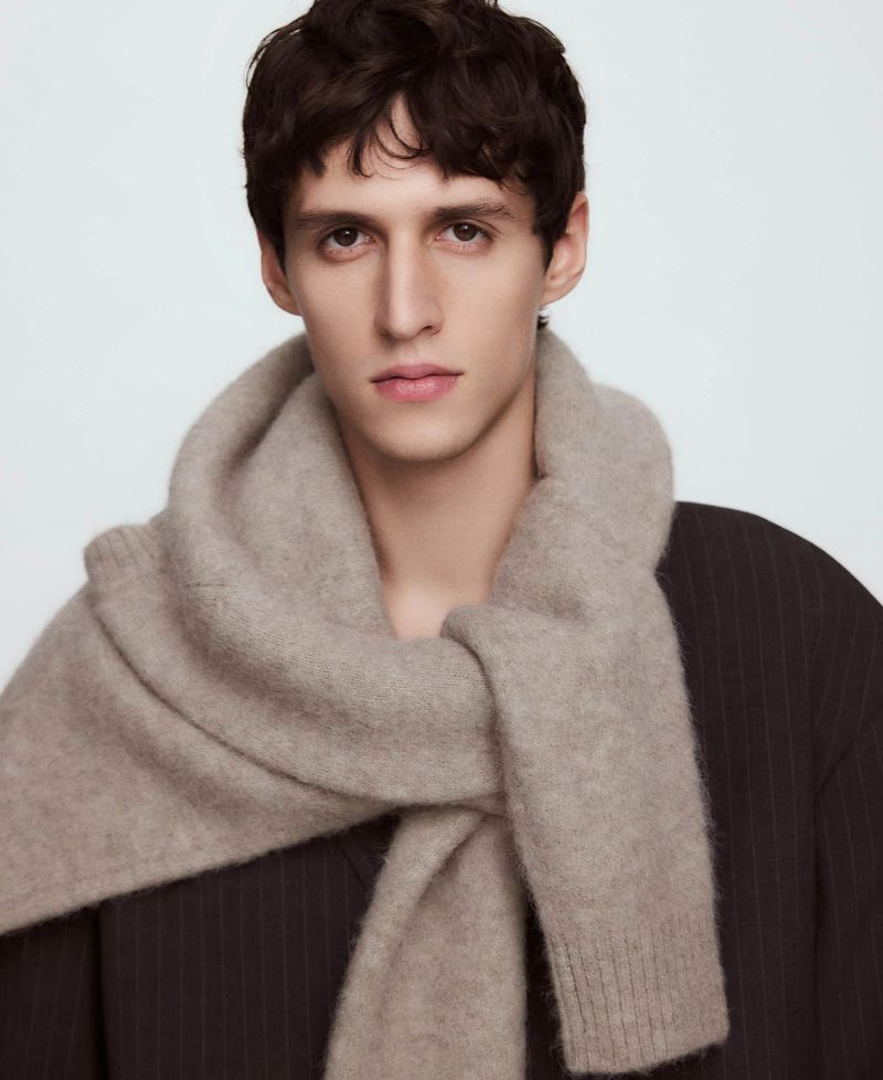 Portrait of a dark-haired male model wearing a neutral-colored scarf and pinstripe jacket against a soft white background. Fashion editorial.