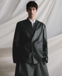 Model wearing a black jacket and white collared shirt against a draped fabric backdrop.