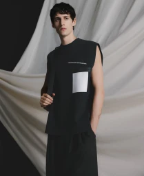 Male model wearing a black sleeveless top with white pocket detail against draped white fabric background