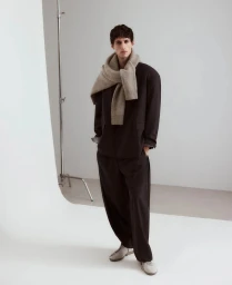 Model wearing a dark oversized suit and beige knit sweater draped over shoulders in a white studio setting.