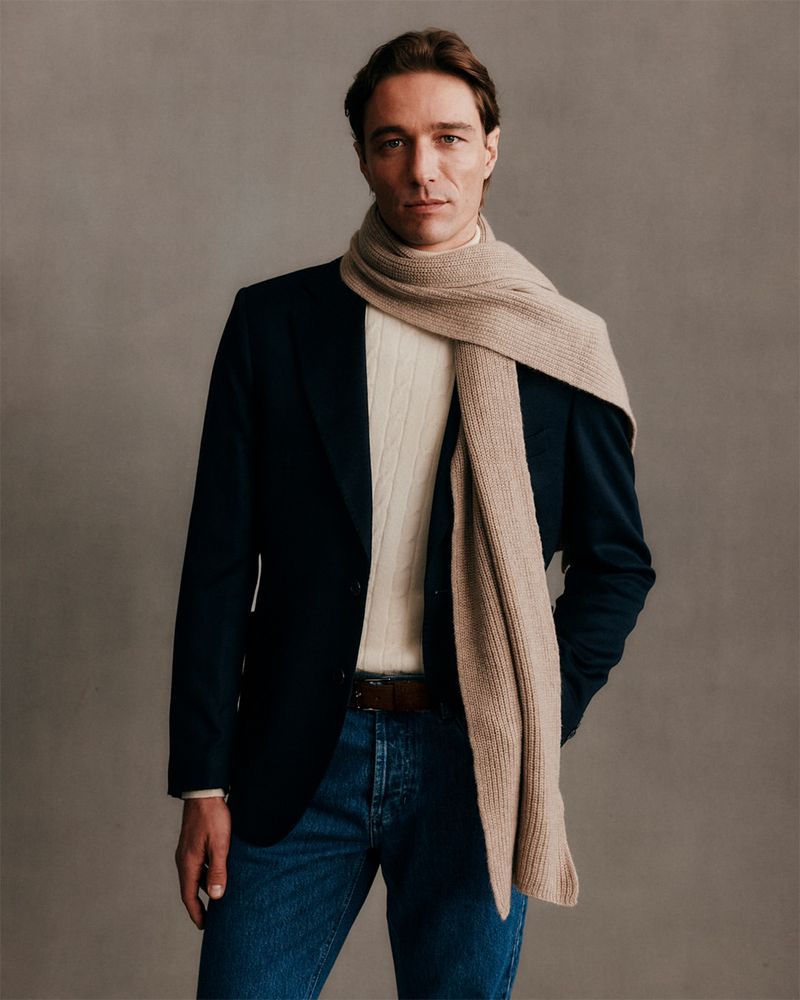 Man in navy blazer, white sweater, jeans, and beige scarf stands against gray background. Fashionable winter outfit.