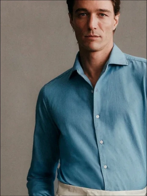 Close-up of an adult male wearing a light blue button-down shirt against a neutral background in a studio setting.