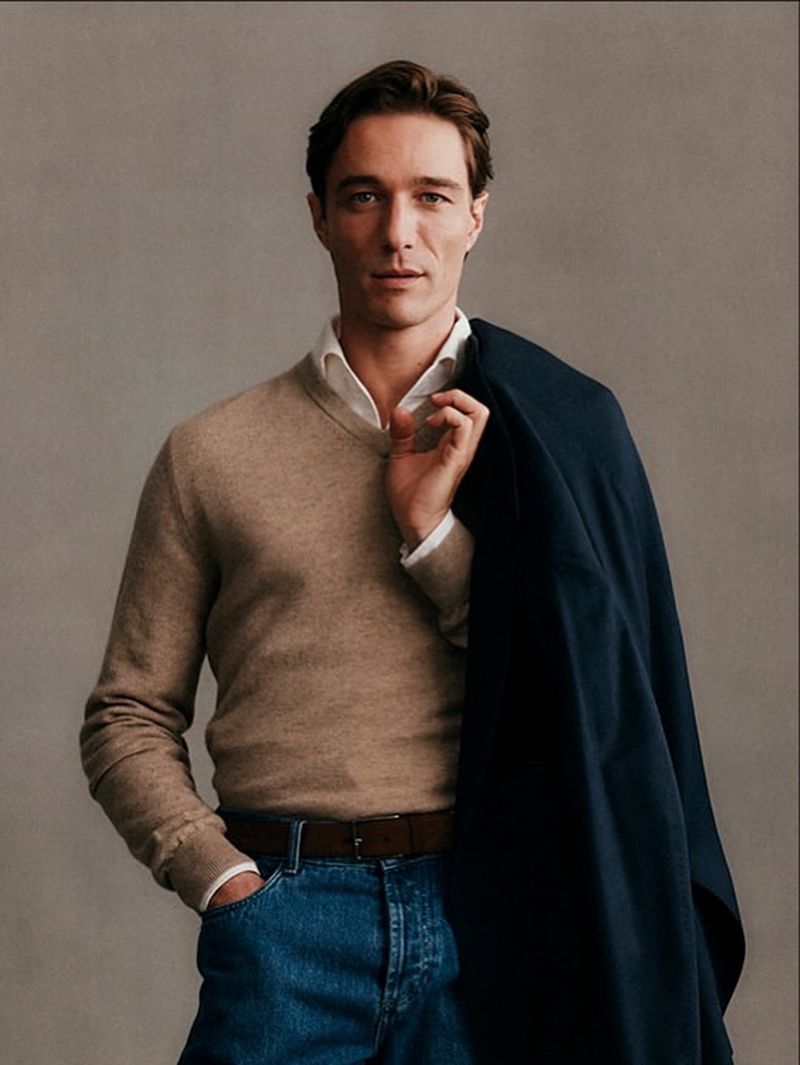 Model wearing beige V-neck sweater, white shirt, and denim jeans while holding dark jacket over shoulder against grey wall