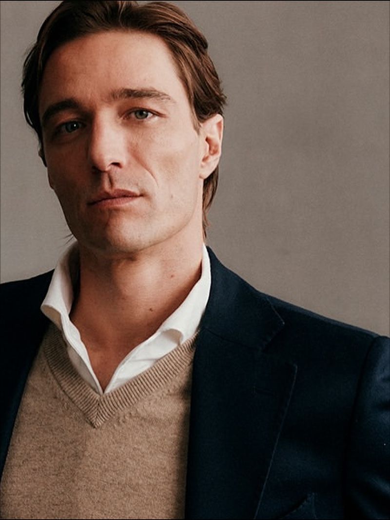 Portrait of an adult male wearing a white shirt, tan sweater, and navy blazer. Head and shoulders view on grey studio background.