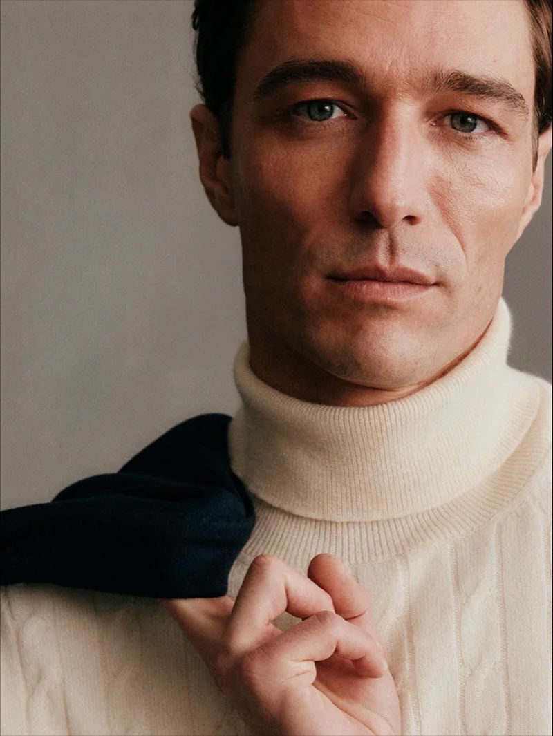 Close-up portrait of a man wearing a cream cable-knit turtleneck sweater with a dark navy garment draped over his shoulder.