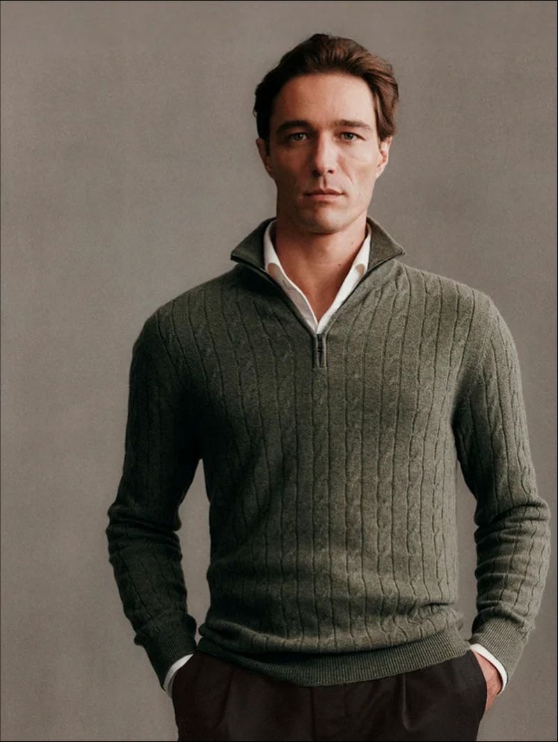 Man wearing a green cable-knit quarter-zip sweater over a white collared shirt against a plain grey studio background.