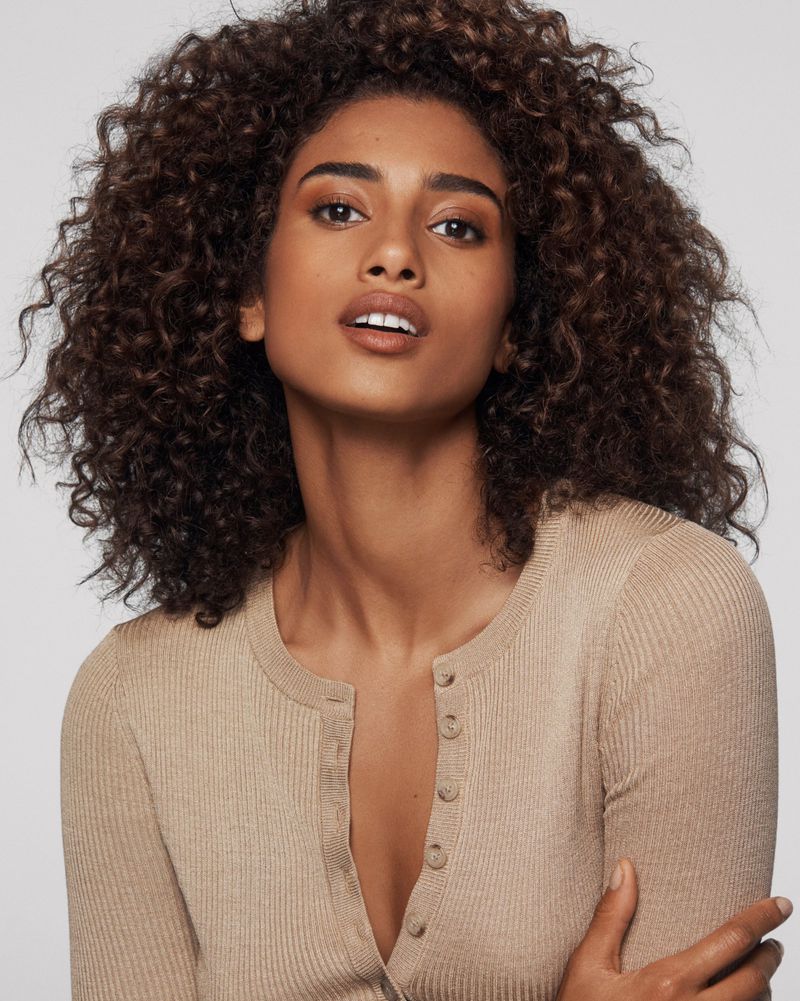 Close-up studio shot of a beautiful adult woman with curly brown hair, tan complexion, wearing a beige top against a neutral background.