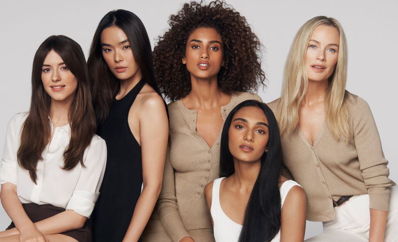 Five diverse women pose in a studio shot, wearing casual tops. Two women sit in front; three women stand behind them. The background is plain.