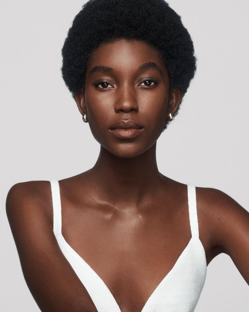 Close-up studio portrait of a young adult Black female model wearing a white top, posing against a neutral grey background.