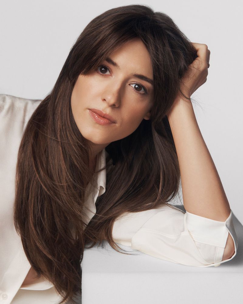 Portrait of a brunette woman with long hair and a white blouse leaning on a white surface in a studio setting on a white background.