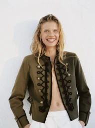 Model wearing an olive green military-style jacket with black braiding and white trousers against a plain white wall