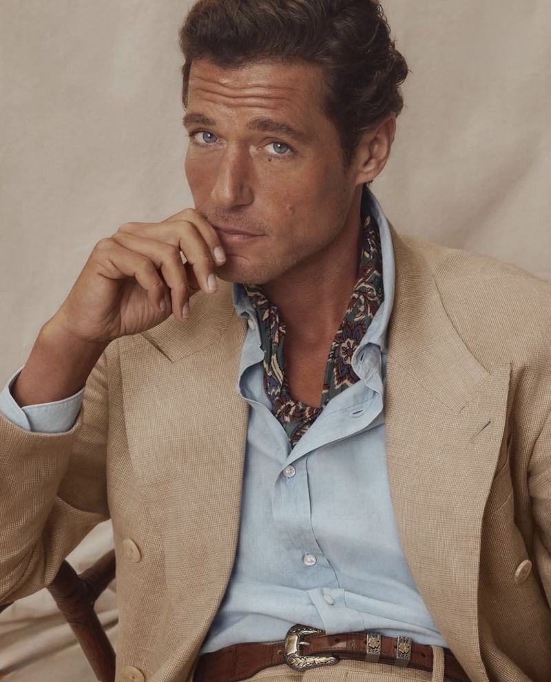 Portrait of a male model in a tan suit jacket, light blue shirt, and printed ascot, posing in a photo studio setting with neutral backdrop.