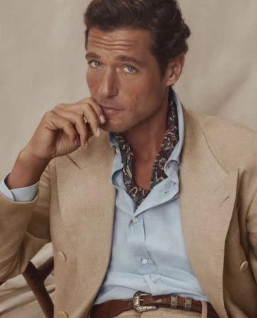 Portrait of a male model in a tan suit jacket, light blue shirt, and printed ascot, posing in a photo studio setting with neutral backdrop.