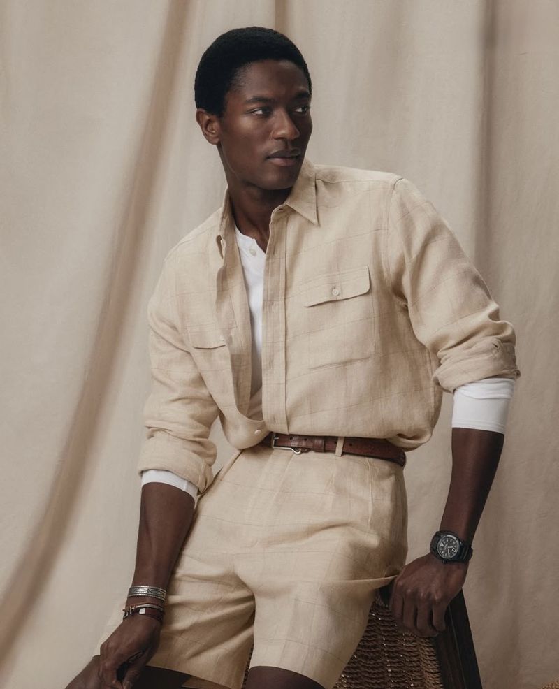 A stylish Black male model poses in a light beige linen shirt and shorts with a brown belt, accessorized with bracelets and a watch.