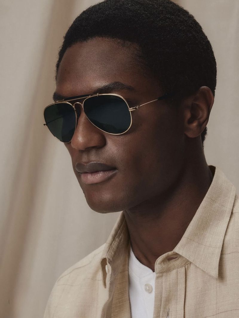 Close-up of a black man wearing aviator sunglasses and a light-colored collared shirt against a neutral background. Fashion portrait.