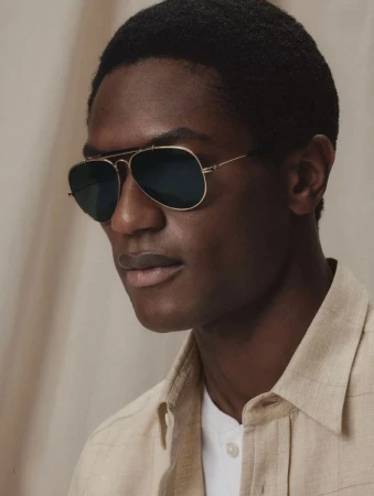 Close-up portrait of a black man wearing dark aviator sunglasses and a light linen shirt against a neutral textured backdrop.